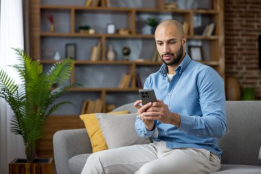 Bir adam rahat bir kanepede rahat bir şekilde oturup, modern bir oturma odasında modern eğlence, teknoloji ve sosyal medya bağlantısının tadını çıkarırken akıllı telefonuyla izliyor ve kaydırıyor.