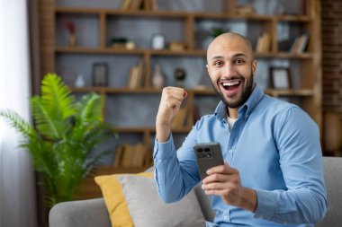 Evdeki kanepede oturan mutlu genç adam, iyi haberler alıyor ve akıllı telefonuna parlak bir gülümseme ve kaldırılmış bir yumrukla bakarken başarısını kutluyor.