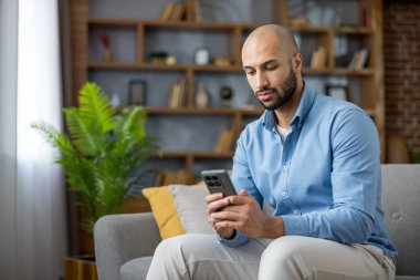 Akıllı telefonuna odaklanan genç kel bir adam, modern gri bir kanepede oturmuş, sosyal medyayla ya da internet iletişimiyle meşgul.