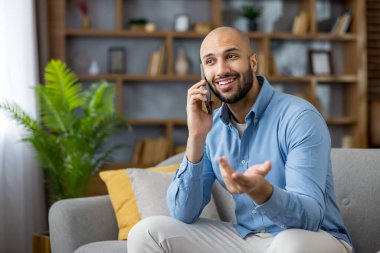Zeki bir telefonda gülümseyen ve sohbet eden kel bir adam. Parlak bir oturma odasındaki modern bir koltukta rahatlarken. Arkadaşça bir video ya da sesli arama sırasında rahat ve kendinden emin.