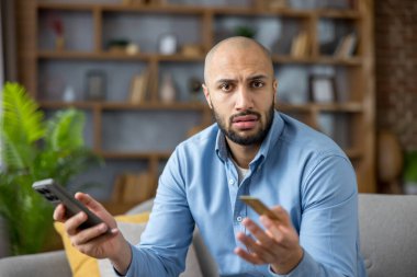 Evde elinde akıllı telefon ve kredi kartı olan kel bir adam, çevrimiçi ödeme hatası veya mobil alım sırasında dolandırıcılık şüphesiyle hayal kırıklığına uğramış, endişeli ve güvenlik konusunda endişeli.