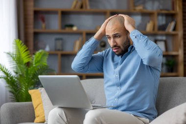 Evde çalışan genç adam, dizüstü bilgisayarında şok ve hüsranla tepki veriyor. Bir hata ya da kötü haber üzerine kafasını tutarken oturma odasında stres yapıyor ve endişeleniyor.