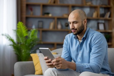 Genç adam evdeki rahat, modern bir kanepeye yaslanmış, yerel oturma odasındaki internet uygulamaları, haberler veya sosyal medya-günlük, bağlı boş vakitler için bir tablete odaklanmış.