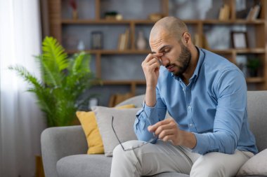 Yorgun ve bunalmış genç bir adam evdeki bir kanepede oturur. Yorgunluktan ve stresten gözlerini ovuşturur. Yorgunluk ve hayal kırıklığını gösterir.