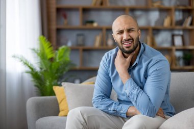 30 'lu yaşlarında bir adam evinde bir kanepede oturuyor, boğazı ağrıdığı için kaşlarını çatıyor ve boynuna dokunuyor, kötü görünüyor ve soğuk algınlığı veya bademcik iltihabı belirtileri gösteriyor.