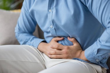 Hazımsızlık ve kramplardan dolayı karın bölgesini sımsıkı tutan, günlük kıyafetlerle evde oturan, ağrı, hastalık ve sindirim sıkıntısı gösteren bir adam.