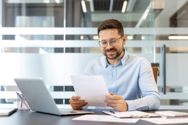 Bir belgeyi incelemeye odaklanan mutlu işadamı, çağdaş bir şirket ortamında dizüstü bilgisayar ve evrak işleriyle dolu bir masada oturuyor, başarı ve büyümeyi temsil ediyor.