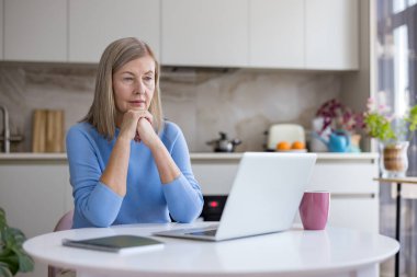 Üst düzey bir kadın evde çalışırken ya da ders çalışırken düşünceli bir şekilde bilgisayarını parlak, modern bir mutfakta kullanarak internete bağlanıyor ve fikirleri yansıtıyor.