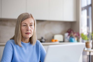 Olgun bir kadın, parlak, modern bir mutfakta dizüstü bilgisayara odaklanmış, uzaktan çalışıyor ve internette geziniyor, aktif üst düzey yaşam, dijital beceriler, bağımsızlık ve konforu gösteriyor