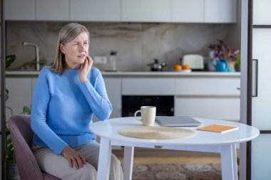 Son sınıf öğrencisi diş ağrısı ve çene ağrısı çekiyor. Mutfak masasında eli yanağında oturuyor. Rahatsızlığını dile getiriyor ve acilen diş bakımına ihtiyacı var.