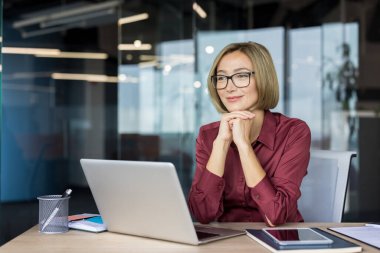 Modern ofiste gözlüklü bir iş kadını, dizüstü bilgisayar kullanıyor ve gülümsüyor, başarılı iş stratejileri ve gelecekteki büyüme fırsatlarını yansıtıyor.