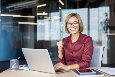Modern ofis masasındaki iş kadını, başarıyı kutluyor, bir dizüstü bilgisayar üzerinde çalışırken gülümsüyor ve yumruğunu kaldırıyor, başarıyı, güveni ve profesyonel zaferi iletiyor.