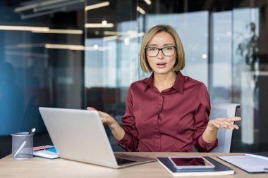 İş kadını elinde dizüstü bilgisayarla masasında oturuyor, elleri açık omuz silkiyor, modern bir ofiste çalışırken kafa karışıklığı ve hayal kırıklığını iletiyor.