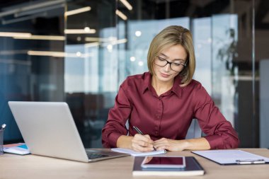 Masasında çalışan gözlüklü bir iş kadını, bir not defterine notlar yazmaya odaklanıyor, çağdaş bir şirket ortamında dizüstü bilgisayar ve çeşitli ofis malzemeleri kullanıyor.