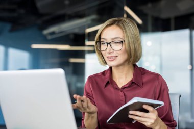Gözlüklü deneyimli bir iş kadını çevrimiçi video görüşmesine katılıyor, elinde bir defter tutarken konuşuyor ve gülümsüyor ve modern ofis ortamında dizüstü bilgisayarının başında çalışıyor.