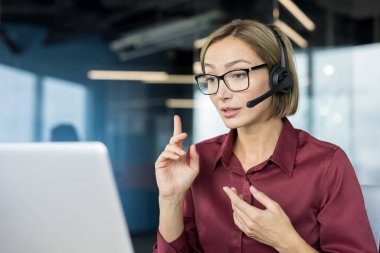 Profesyonel bir kadın kulaklık ve gözlük takıyor, aktif olarak konuşuyor ve el kol hareketi yapıyor, bir ofis ortamından online müşteri hizmetleri veya teknik destek sağlarken, müşterilere yardım etmeye odaklanıyor.