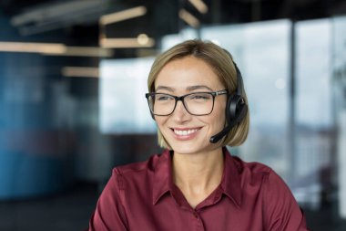 Gözlüklü ve kulaklıklı gülümseyen kadın modern bir ofiste profesyonel bir çağrı merkezi ajanı olarak çalışırken kendine güvenen ve arkadaş canlısı bir şekilde müşteri desteği ve yardım sağlıyor.