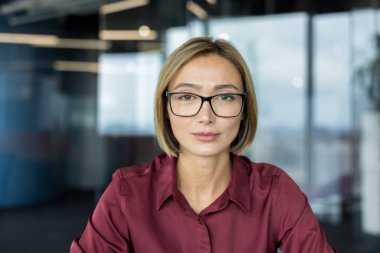 Kurumsal kadın, kısa sarı saçlı ve gözlüklü bulanık ofis ortamında doğrudan izleyiciye bakıyor, özgüven ve profesyonelliği ifade ediyor.