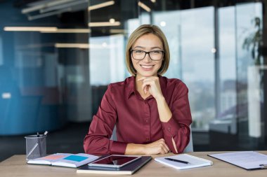 Asyalı iş kadını gülümsüyor ve kendinden emin görünüyor, elinde tablet ve defterle modern bir ofis masasında oturuyor, kurumsal bir ortamda profesyonelliği ve başarıyı temsil ediyor.