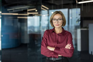 Gözlüklü, kestane rengi gömlekli, kollarını kavuşturup kameraya kararlı bir ifadeyle bakan, modern bir ofiste duran kendine güvenen bir iş kadını.