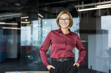 Gözlüklü, bordo tişörtlü, elleri ceplerinde gülümseyen, çağdaş bir iş yerinde duran genç bir bayan profesyonel.