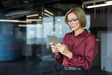 Gözlüklü genç iş kadını dijital tablet üzerinde araştırma yapıyor, cam duvarları olan çağdaş bir yaratıcı ofiste duruyor, profesyonel çalışmalarda teknoloji entegrasyonunu temsil ediyor.