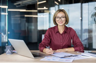 Gözlüklü ve kırmızı tişörtlü genç bir iş kadını modern ofis masasında bilgisayarlı, tabletli ve grafikli evrak işleriyle uğraşıyor.