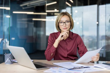 Asyalı iş kadını masada oturuyor, elinde bir dizüstü bilgisayar ve finansal çizelge ile çevrili profesyonel bir ofis ortamında elinde kağıtlarla kameraya bakıyor.