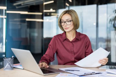 Gözlüklü iş kadını dikkatle bir dizüstü bilgisayarda çalışıyor ve ofis masasındaki belgeleri inceliyor, odak noktasını, şirket verimliliğini ve modern işyeri verimliliğini temsil ediyor.