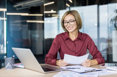 İş kadını ofis masasında oturuyor, gülümserken bir belge okuyor, mesleki işi ve kurumsal görevlerinde başarı ve başarı buluyor.