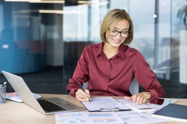 Hesap makinesiyle çalışan gülümseyen iş kadını, finansal belgelere yazı yazan, ve modern bir ofis masasında dizüstü bilgisayar kullanan, odaklanmış ve verimli bir profesyonel faaliyet gösteren