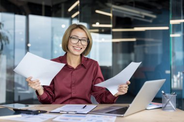 Gülümseyen Asyalı iş kadını iş belgelerini değerlendiriyor, profesyonel başarının tadını çıkarıyor ve modern ofis ortamında masasında güvenle çalışıyor.