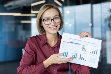 Genç iş kadını gülümsüyor, gözlük takıyor ve elinde gelir-zarar tablosu karşılaştırma tablosu olan bir belge tutuyor.