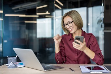 İş kadını masasında başarıyı kutluyor, dizüstü bilgisayar üzerinde çalışırken akıllı telefon tebligatına gülümsüyor, profesyonel zafer, çevrimiçi galibiyet ve işyeri başarısını iletiyor