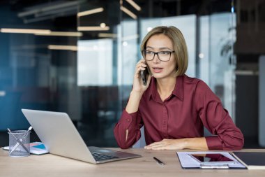 Modern ofisteki Asyalı iş kadını, masa başında çalışırken cep telefonuyla konuşuyor, müşteri iletişimine, görevleri yönetmeye ve dijital işbirliğine odaklanıyor