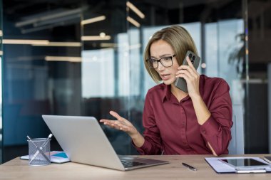 Stresli iş kadını gözlük takıyor. Bilgisayarına bakarken zor bir telefon görüşmesi yapıyor. Sorunlarla başa çıkıyor ve modern ofis işyerinde kötü haberler alıyor.