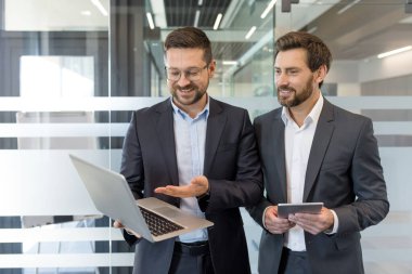 Parlak bir ofis ortamında işbirliği yapan, fikirleri paylaşan ve bir dizüstü bilgisayar ve dijital tablet üzerindeki bilgileri inceleyen, takım çalışmalarını ve modern iletişimi vurgulayan iki güler yüzlü işadamı