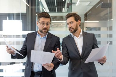 İki iş arkadaşı, bir kurumsal ofis ortamında strateji ve karar verme konusunda işbirliği yaparak önemli proje belgelerini inceliyor ve tartışıyor.