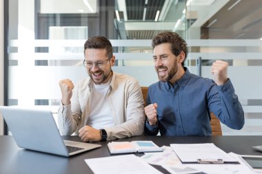 İki işadamı büyük bir dönüm noktasını kutluyor. Modern bir ofiste dizüstü bilgisayarın başında sevinçle gülüyorlar. Heyecanlı gülümsemeleri, takım çalışmalarını ve muzaffer iyi haberleri paylaşıyorlar.