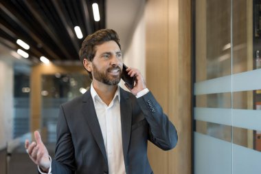Kendine güveni tam bir iş adamı akıllı telefonuyla neşeli bir konuşma yapıyor. Parlak, modern ofis koridorunda yürürken elini sallıyor ve gülümsüyor.