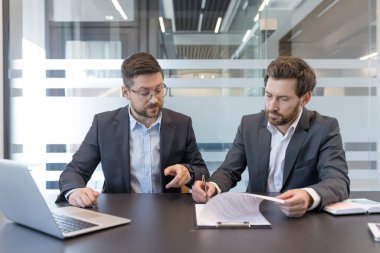 Karanlık bir ofis masasında oturan iki işadamı, önemli bir toplantıda bir dizüstü bilgisayar ve belgelerle birlikte yasal bir sözleşmeyi gözden geçiriyorlar ve imzalıyorlar.