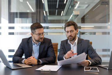 Modern bir konferans odasında üç işadamı finansal raporları analiz eder ve birlikte dizüstü bilgisayarlar, tabletler ve evraklar üzerinde strateji oluşturur.