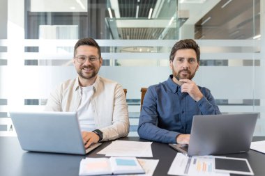 İki profesyonel iş adamı gülümsüyor, dizüstü bilgisayarlarıyla konferans masasında oturuyor, çağdaş bir ofis ortamında verimli bir takım çalışması yapıyorlar.