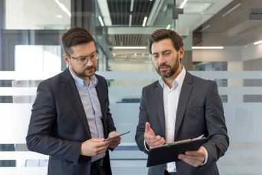 Modern bir ofiste işbirliği yapan ve belgeleri analiz eden iki profesyonel işadamı, stratejik iş geliştirme ve ortaklık üzerine odaklanmış bir tartışmaya giriştiler.