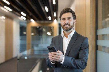 Profesyonel işadamı takım elbise giyiyor ve gülümsüyor, çağdaş bir ofis koridorunda duruyor ve bir akıllı telefonla iletişim ve bağlantıyı temsil ediyor.
