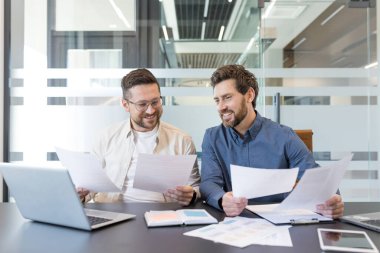 Modern ofis toplantı odasında gülümseyen ve iş belgelerini gözden geçiren iki erkek iş arkadaşı dizüstü bilgisayarı ve masa başında evrak işleriyle birlikte çalışıyorlar.