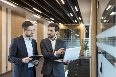 İki profesyonel iş adamı işbirliği yapıyor ve fikirlerini paylaşıyor, biri elinde bir dizüstü bilgisayar, diğeri ise bir pano tutuyor, modern ofis koridorunda önemli iş konularını tartışıyorlar.