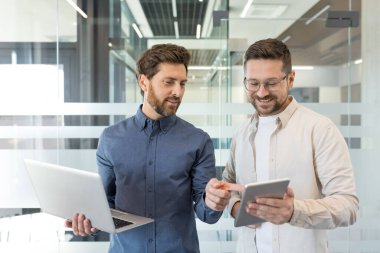 İki gülümseyen sakallı iş adamı modern bir ofiste duruyor, biri dizüstü bilgisayar tutarken diğeri dijital tablet ekranı gösteriyor, paylaşılan bir projede aktif olarak işbirliği yapıyorlar.