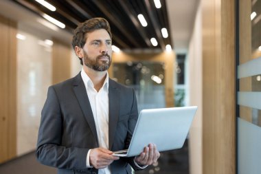 Çağdaş ofis koridorunda duran kendine güvenen bir iş adamı. Bir dizüstü bilgisayar tutmak ve dikkatle başka yere bakmak. Profesyonelliği somutlaştırıyor. Başarı. Ve kurumsal bir ortamda stratejik düşünme