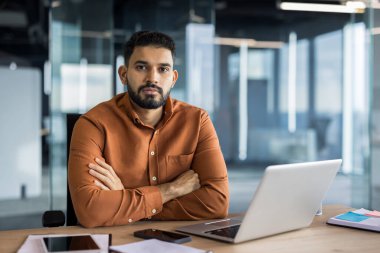 Modern bir ofisteki kendine güvenen işadamı masasında oturuyor, kollarını kavuşturmuş, ciddi görünüyor ve odaklanmış bir şekilde. Etrafı dokümanlar ve iş eşyalarıyla çevrili bir dizüstü bilgisayar üzerinde çalışıyor.